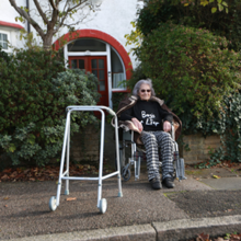 An old woman in a wheelchair on a sidewalk in front of a house wearing a black shirt with the text "Boy's a Liar"
