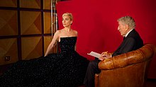 A photograph of Bennett sitting in an armchair sketching a portrait of Gaga, who is stood beside him waring a black dress.