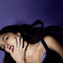 Olivia Rodrigo in a black dress biting her thumb on a dark purple floor. Her four rings spell out G–U–T–S