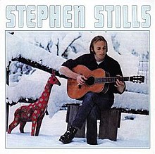 Stephen Stills sitting outside in the snow and playing a guitar, with a toy giraffe nearby