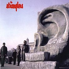 The band members standing near a large sculpture of a ear behind a building. The album's title is engraved in large blocks of stone.