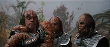 Two aliens watch their leader fight a slimy worm that has wrapped itself around his arm and neck. The aliens are wearing metallic armor with decorative script on their gauntlets; their hair is long and black, and their foreheads are bumpy and ridged.