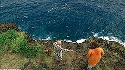 A cliffside by the sea. A man in plaid pajamas talks to another wearing an orange shirt.