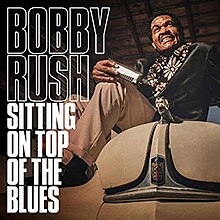 Bobby Rush, sitting on the hood of a vintage car and holding a harmonica