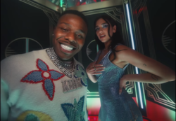In an art-deco style elevator, DaBaby smiles at the camera wearing a white outfit with multi-coloured designs, while Dua Lipa, also looking at the camera, touches her chest and puts her hand on her hip, wearing a blue dress.