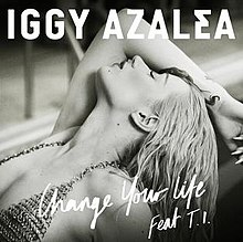 A black-and-white portrait of a young woman with wet, blond hair wearing a chain-mail bikini striking a dramatic pose. Centred across in white cursive font stands "Change Your Life Feat. T.I". Above the woman in white bold font stands the name "Iggy Azalea".