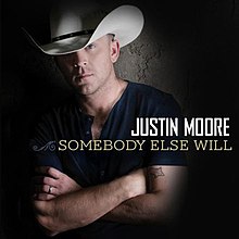 The cover features Moore wearing a navy blue shirt and a white cowboy hat with a black ring around it, standing behind a concrete wall with his arms crossed. The artist's name and song title are colored white and beige respectively.