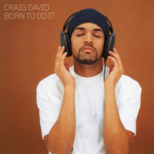 David standing against a brown background wearing a dark beanie and headphones with his eyes closed and his hands touching his headphones.