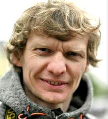 Colour press photo of smiling, clean-shaven man with wavy blond hair.