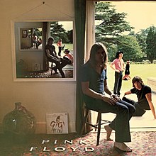 A room with the doors open with the outside showing trees and a grass field with one person sitting in a chair and the rest of the band outside. The text reading "PINK FLOYD" is placed on the floor. On the floor, a vase and a vinyl album of Gigi leaning against the floor. Hanging on the wall is a looping image of the same scene but with the persons in different positions.