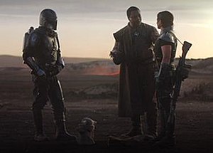 Four protagonists from The Mandalorian stand on rocky terrain, with volcanic embers and an evening horizon visible behind them.