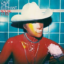 A photo of a man in a white cowboy hat and red rubber shirt with a bronze-colored face and a yellow streak going down the side of his face, stood in front of a blue tile wall.