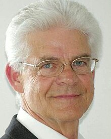 Publicity shot of smiling, clean-shaven man with gold-rimmed spectacles and white hair.