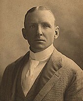 Portrait of a man in a jacket with a dark collar wearing a white shirt and tie.