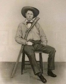 Sepia-tone photograph of a young man sitting on a chair against a plain wall. He has a narrow face and is clean-shaven, and wears a broad-rimmed hat, bow tie, and loose-fitting shirt and pants. He sits leaning back, with his legs pushed out in front of him. His right hand is bandaged, and in his right arm he cradles a large rifle, with its stock near his face and the barrel resting on the floor.