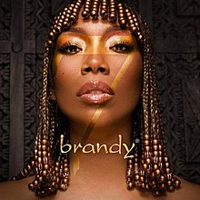 A black woman stands in front of a carved wooden background. She wears her hair with micro braids and a number of golden beads that cover her fringe and the ends of her braids. Across her first is the number 7 in gold lettering and beneath is the word "brandy".