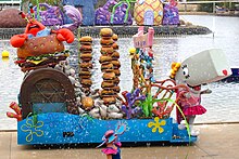 A costumed mascot of Pearl and an animatronic Mr. Krabs standing on a parade float