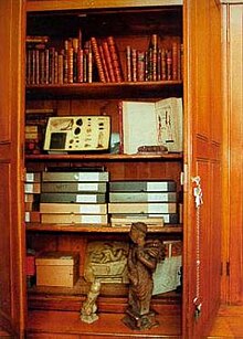 The inside of a wooden cupboard with four shelves. The top two shelves have books (two are open); the third shelf down has carboard boxes (some labelled "WITT") and the bottom shelf has a cardboard box with three sexually graphic statues