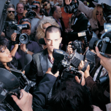 Robbie Williams stands surrounded by a group of photographers and interviewers attempting to interview him or take a picture of him.