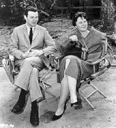 A black and white photograph of Alan J. Pakula seated next to Harper Lee in director's chairs watching the filming of To Kill a Mockingbird