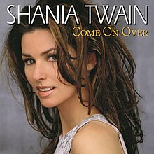 Photograph of a woman standing in front of a grey background. She is half smiling and wearing a sleeveless silver gown. The words SHANIA TWAIN are written at the top of the image in white capital letters. The words COME ON OVER are written below in gold capitals.