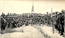 A black and white photo of US Army soldiers on 3 July 1898, in an unside down V type formation on top of Kettle Hill, two American flags in center and right. Soldiers facing camera.