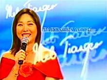 An image of Mel Tiangco smiling, wearing a red top and holding a microphone, on a blue backdrop. The series title is displayed on the center of the image.