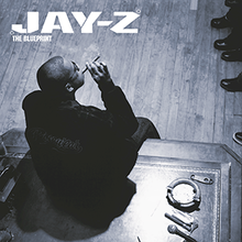 Jay-Z sitting on top of a table smoking a cigar while wearing glasses and a jacket reading "Rocawear" at the back. On the table, an ashtray a pack of cigars, microphone, and other items. Out-of-background, people are seen, with the shoes only viewable.