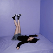 Olivia Rodrigo lying on a bed with her legs raised. The bed and walls of the place are purple.