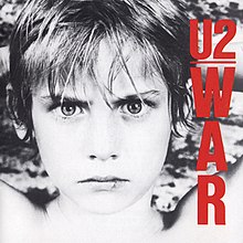 A black and white image of a young boy with his hands behind his head staring fiercely at the viewer. On the right side in red text, the band name is horizontally arranged and underlined, with the album title vertically stacked underneath.