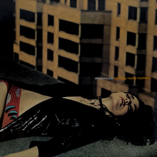 Madonna laying down at the corner of a rooftop, with yellow buildings present behind her. She wears a black shining jacket.