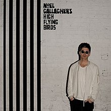 Noel Gallagher standing on the right side in front of a white brick wall with black stripes to the left of him, and Noel Gallagher's High Flying Birds written above him.