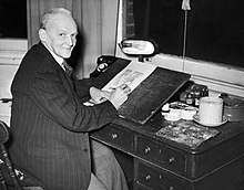 Black and white image of a man wearing a suit, seated in front of a desk.