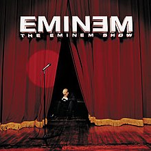 Image of a stage with a microphone stand and a spotlight pointing to where a performer would be. There is no performer seen. Curtains are slightly opened to reveal a man seated on a low chair. He is wearing a suit and his head is facing down. His hands are on his mouth to indicate worry. At the top, sits a 3D version of the Eminem logotype. Below it is text that reads "The eminem show" in a smaller font
