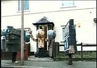 A person dressed in a cardboard costume with a large cardboard head and arms exits the central door of a two storey house which is painted cream. A short path leads directly from the door through a small garden to the pavement outside. To the left of the path a man dressed as a camcorder stands on the pavement behind a lamppost, facing the doorway. At the very end of the path, to the right, a man dressed as a microphone stands sideways, looking at the doorway. On the pavement to the right of the path a person dressed as a clapperboard stands looking down the pavement towards the person dressed as a camcorder.