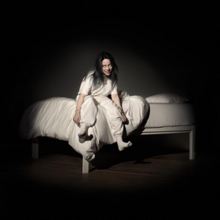Eilish sits on the edge of a white bed, in front of a dark background. She wears white clothing, with white eyes while smiling demonically at the camera.