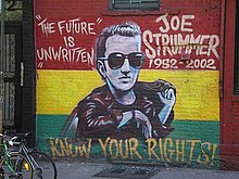 A photograph of the painting of the memorial mural of Joe Strummer on the wall of the Niagara Bar in the East Village in New York City. The mural depicts Strummer (centre) surrounded by the words "THE FUTURE IS UNWRITTEN" (on the left), "JOE STRUMMER 1952–2002" (on the right), and "KNOW YOUR RIGHTS!" (bottom) on a horizontal tricolour of red, yellow, and green background