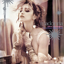 Madonna wearing a wedding dress and heart-shaped earrings is using one of her gloved hands to pull down on her pearl necklace.