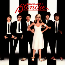 The band members of Blondie standing behind a black and white wall. Debbie Harry (standing center right, with her clenched hands on her hips) is seen wearing a white dress while the rest of the band wears business suits with a black tie.