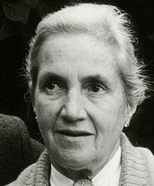 Black and white news photo of an elderly women with an alert expression, no make-up, and white hair tied back, wearing a cardigan.