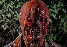 Set against a forest background, a close-up of a skeletal-like face of a man with red blood-like substances dripping from his face and his right eyeball falling out of its socket.