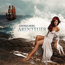 A red-haired woman wearing a long beige dress is sitting on rocks. A wave is hitting the rocks behind her and the words "Andrea Berg Abenteuer" are written on the wave. A pirate ship can be seen in the background in the top left corner.