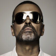A close up of George Michael seen with a white shirt, a beard, and sunglasses, with its lens showing a reflection.