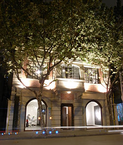 photo of a colonial style building lit up at night. The first two floors of the building are visible, with the top floors obscured by trees with dense foliage. The interior of the gallery can be viewed from outside through two ceiling to floor windows.