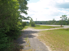 A narrow abandoned road in a wooded area with a two-lane road visible to the right