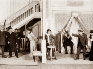 Stage scene in a hotel bedroom, with one man pointing a gun at another, while a woman drags the latter out of the room