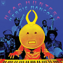 A human figure wearing a horned mask with its face resembling a reel-to-reel tape recorder playing keyboards in the foreground, while four unmasked men in the background hold instruments. The keyboardist has shades of yellow and red, while the musicians in the back blend with the blue background
