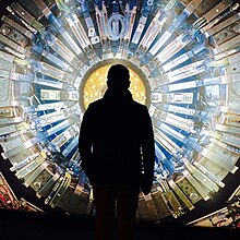 An outline of a man's shadow standing in front of a large metallic circle that is covered in machine parts