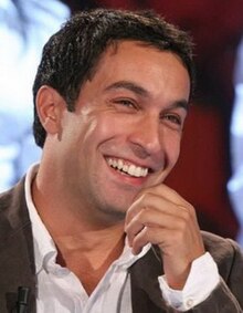 close-up of Pietro Taricone wearing a suit, laughing with his head tilted and one hand held up close to his face, looking just left of camera