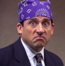 A white man in a suit wears a purple bandana with an exaggerated smug expression on his face.
